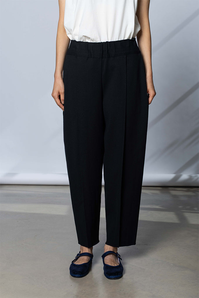 A model wearing Issey Miyake black wide-leg cropped trousers with a gathered elastic waistband, paired with a white top and Drogheria Crivellini navy blue shoes, standing against a light gray studio backdrop.