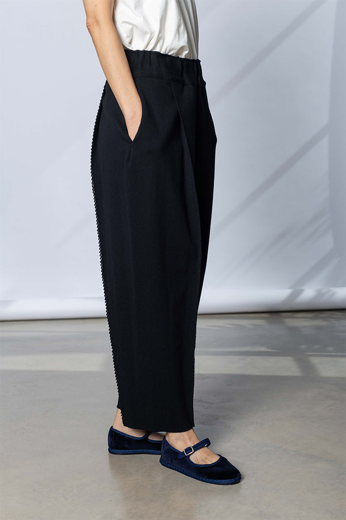 A model is shown from the waist down wearing Issey Miyake black wide-leg trousers with decorative chain detailing along the side seams, paired with a white top and dark navy velvet Mary Jane shoes.