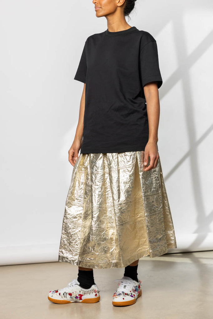 Person wearing a black t-shirt and gold metallic skirt against a white wall.