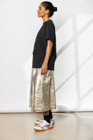 Person wearing a black t-shirt and metallic skirt standing against a white wall.